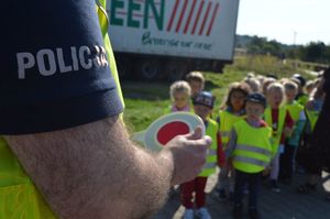 Bok koszulki policjanta z napisem Policja, który w ręku trzyma tarczę do zatrzymywania pojazdów. W tle przedszkolaki w kamizelkach odblaskowych.