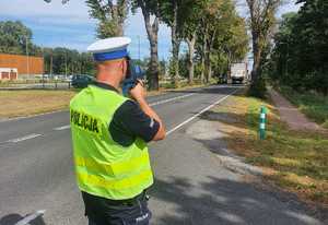 Policjant w mundurze i kamizelce odblaskowej stoi przy drodze trzymając radar prędkości i mierzy nim w kierunku nadjeżdżającego drogą pojazdu osobowego.