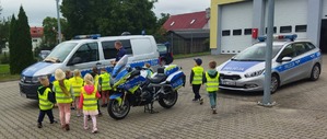 Dzieci w wieku przedszkolnym w kamizelkach odblaskowych stoją przy radiowozie i motocyklu