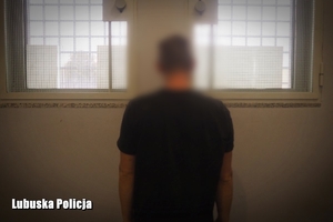 Zatrzymany mężczyzna stoi w policyjnej celi tyłem do zdjęcia na tle okratowanych okien.