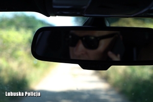 Mężczyzna w okularach przeciwsłonecznych trzyma przy uchu telefon. Jego odbicie widoczne jest w lusterku samochodowym.