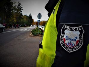 Policjant Wydziału Ruchu Drogowego w odblaskowej kamizelce stoi bokiem do zdjęcia, w tle widać jezdnię oraz cmentarz.