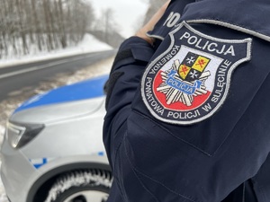 Bok kurtki policjanta z naszywką sulęcińskiej jednostki. W tle radiowóz i zaśnieżona droga.