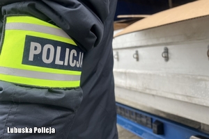 Bok kurtki policjanta z opaską, z napisem Policja na tle zabezpieczonego pojazdu ciężarowego.