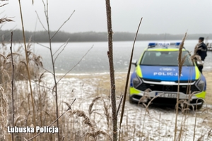 Policjant stoi obok radiowozu na tle jeziora w zimowej aurze.