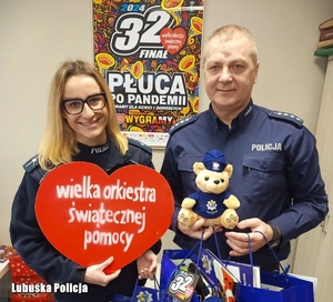 Policjant i policjantka pozują do zdjęcia, trzymając w dłoniach kartonowe serce oraz gadżety przekazane na licytację Wielkiej Orkiestry Świątecznej Pomocy.