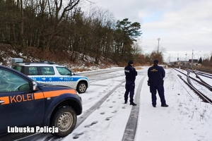 Policjant oraz funkcjonariusz Straży Ochrony Kolei obserwują tory kolejowe. Na zdjęciu widoczne się pojazdy obu służb.