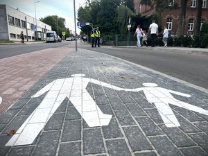 Oznakowanie poziomie droga na pieszych, w tle szkoła i policjanci.