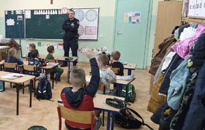 Policjant prowadzi spotkanie edukacyjne dla uczniów na temat bezpiecznych ferii. Znajdują się klasie.