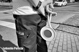 Policjant trzyma w dłoni tarczę do zatrzymywania pojazdów. W tle przejazd kolejowy.