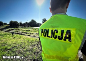Policjant w kamizelce odblaskowej na tle przejazdu kolejowego.