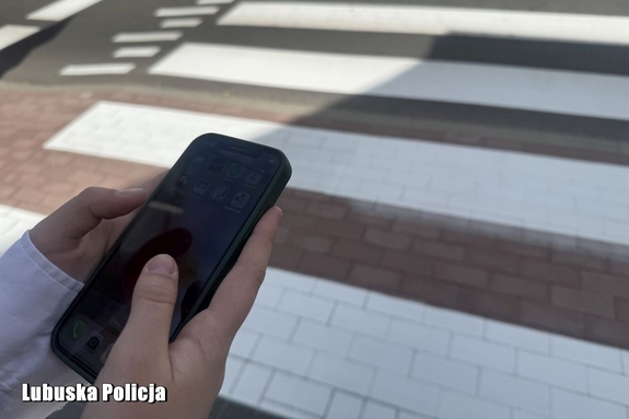 Dziewczynka trzyma w dłoni telefon komórkowy. W tle przejście dla pieszych.